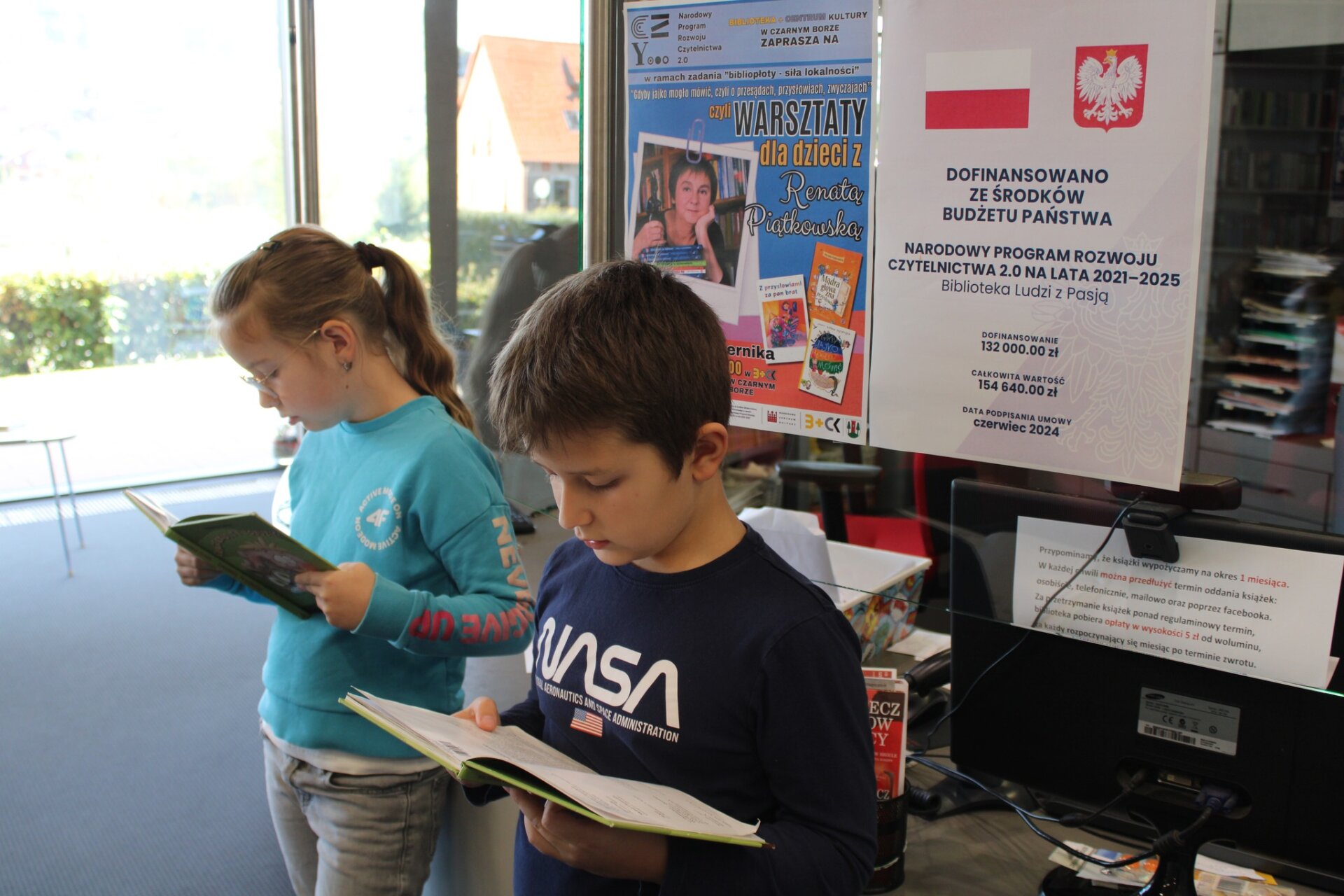 Dwójka dzieci czyta książki za nimi w tle plakaty projektu Blisko "Biblioteka Ludzi z Pasją"