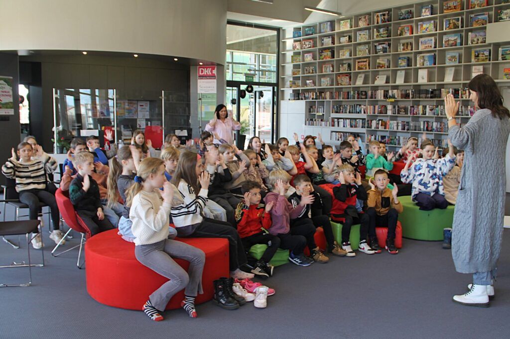 Grupa dzieci w bibliotece podnosi ręce w zabawie z autorka książek, Małgorzata Swędrowską