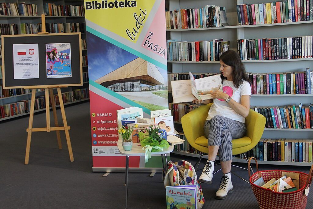 Autorka książek, Małgorzata Swędrowska siedzi na żółtym fotelu w bibliotece i ogląda książkę