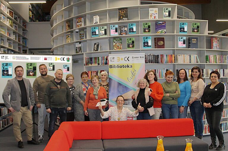 Grupa beneficjentów inicjatyw oddolnych pozuje do zdjęcia w bibliotece na tle baneru BLISKO
