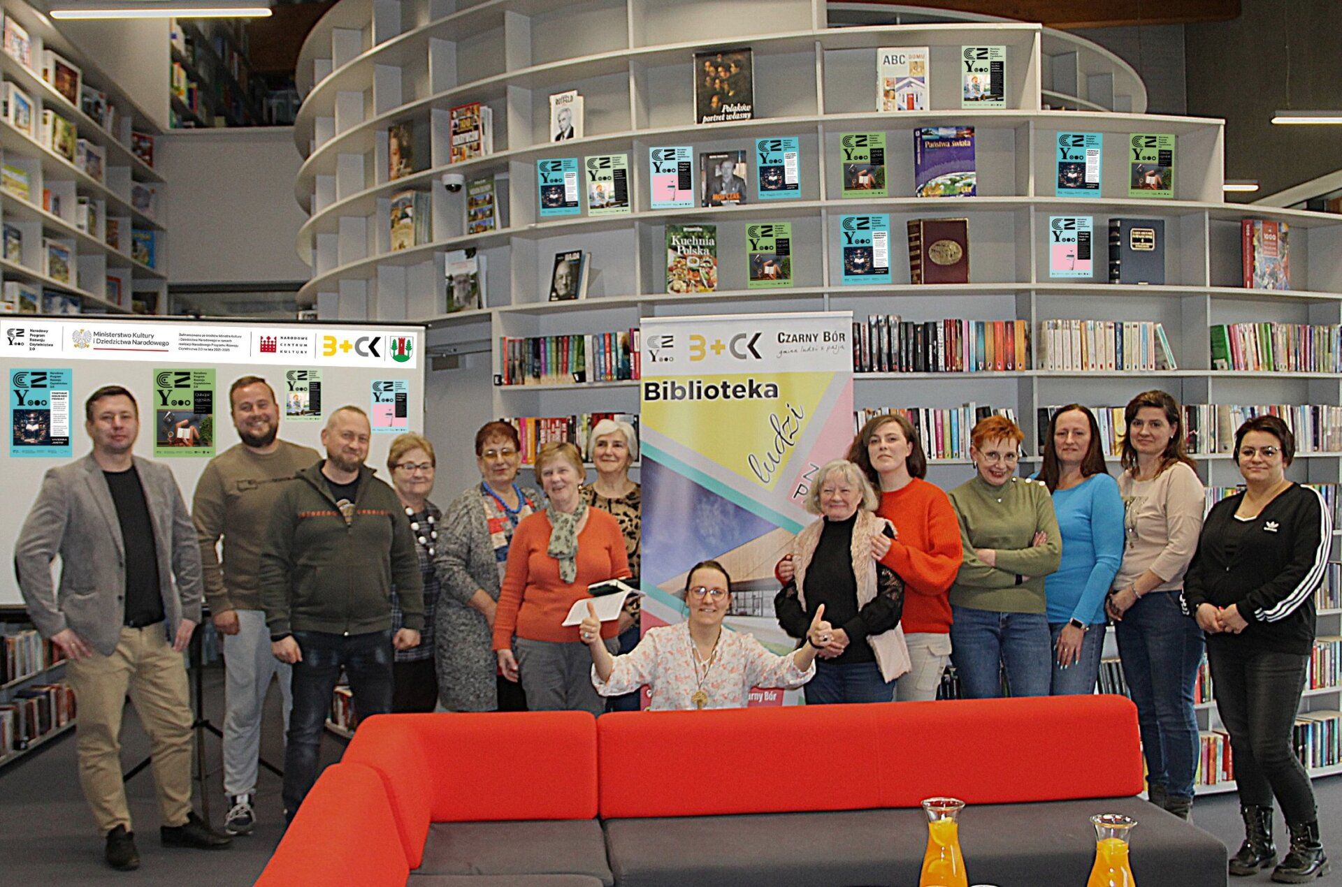 Grupa beneficjentów inicjatyw oddolnych pozuje do zdjęcia w bibliotece na tle baneru BLISKO